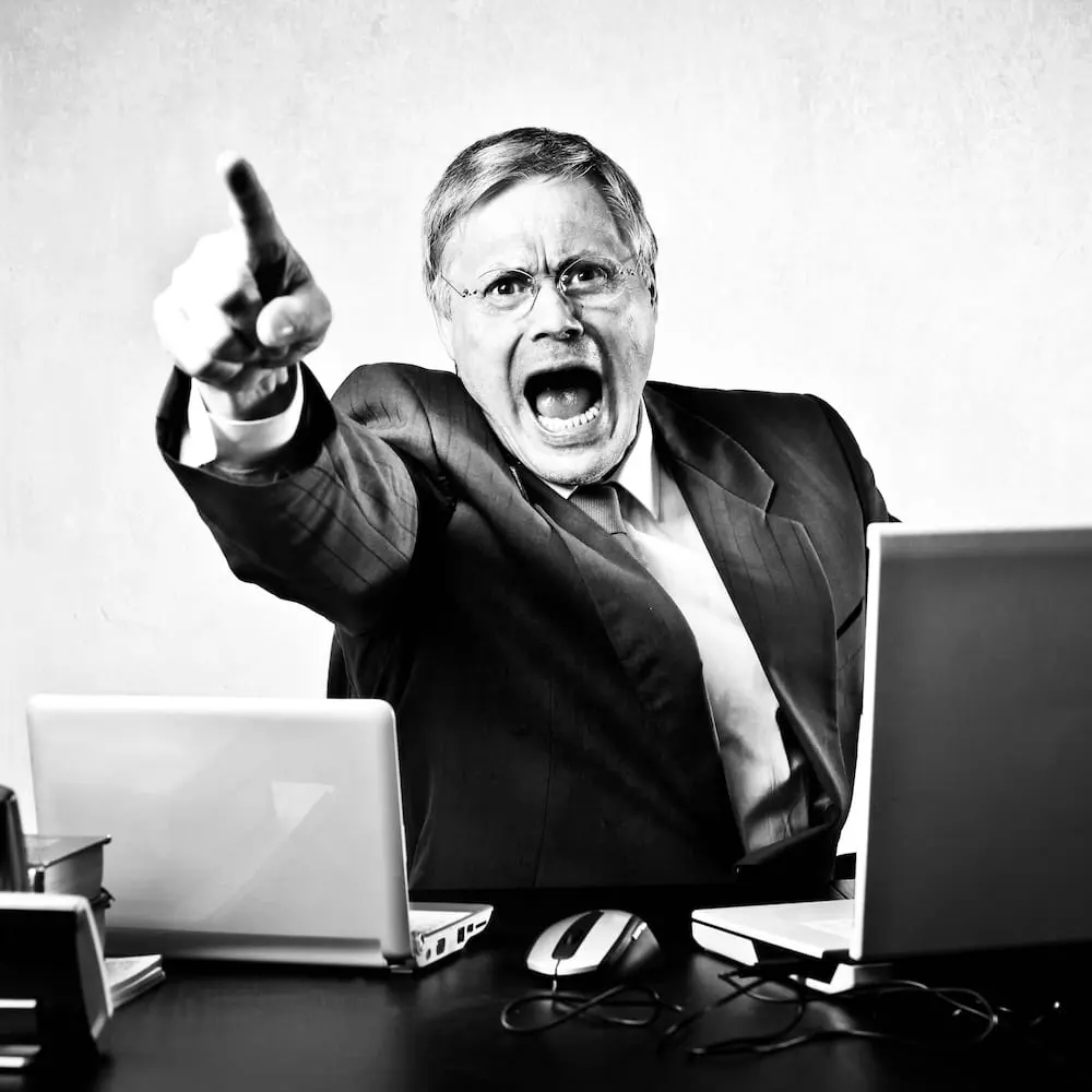 Wenn Überlastung zu Kontrolle, Frustration und Eskalation führt Person sitzt an einem Schreibtisch und zeigt mit ausgestrecktem Finger in den Raum. Das Bild symbolisiert eskalierende Kommunikation, Kontrollverhalten und die Folgen von Überlastung im Führungsalltag.